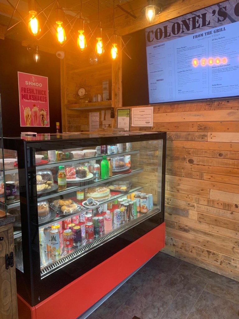 inside Colonel's Grill with wood-clad walls, a digital menu on a screen and a fridge counter with a selection of cakes and canned soft drinks.